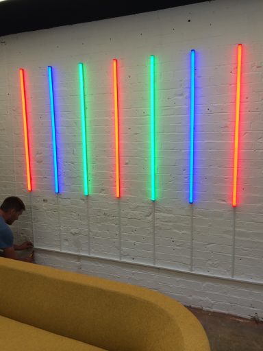 Neon light tubes in red, blue, and green arranged vertically on a white brick wall.