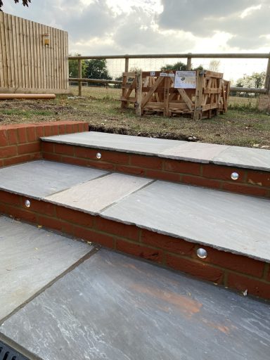 Stone steps leading up to a patio, with a wooden fence and stacked materials in the background.
