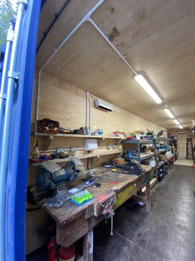 Workshop interior with tools, shelves, and workbench under overhead lighting.