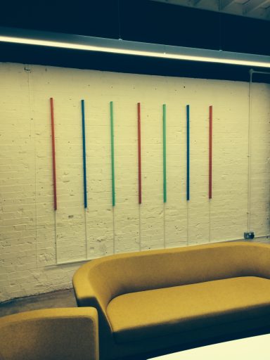 A wall with six vertical colour strips above a yellow couch.