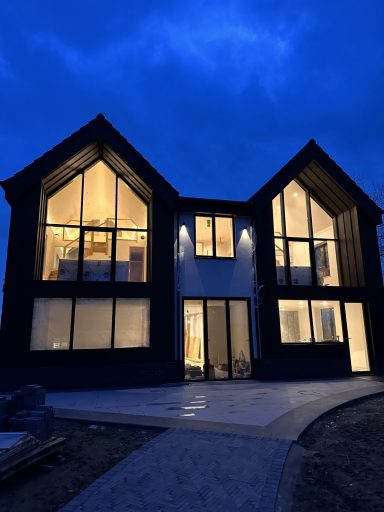 Modern two-storey house with large windows, illuminated at dusk under a cloudy sky.
