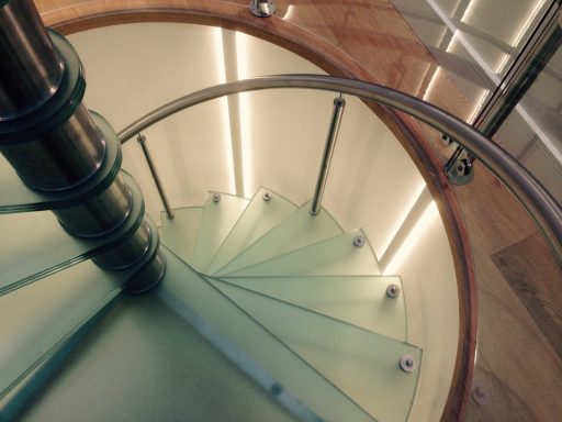 Spiral staircase with glass steps and polished metal railing, softly illuminated.